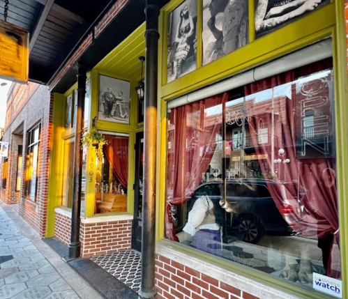 A vibrant storefront with large windows, featuring red curtains and vintage photos, on a brick-lined street.