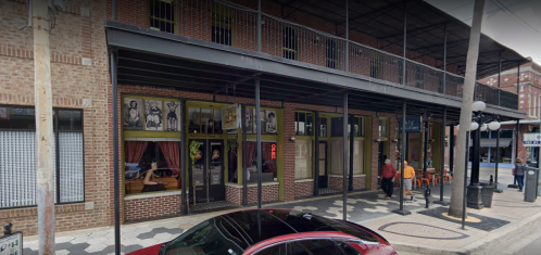 A brick building with a covered porch, featuring large windows and people walking by on the sidewalk.