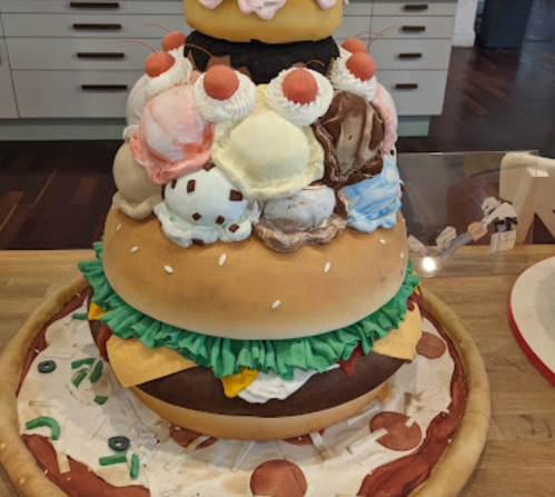 A large, whimsical cake shaped like a towering burger topped with colorful ice cream scoops and decorative toppings.