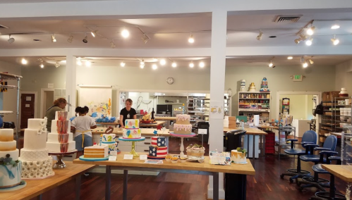 A bright bakery interior with various decorated cakes on display and staff preparing in the background.