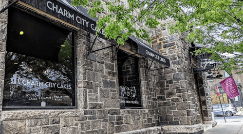 Exterior of Charm City Cakes, featuring stone walls, large windows, and a sign that says "Open." Trees are visible nearby.