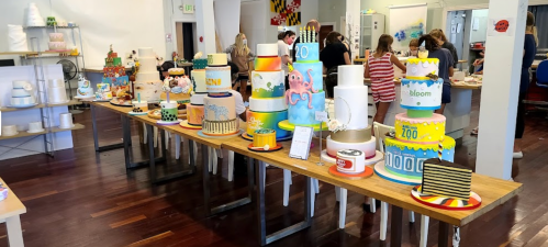 A colorful display of elaborately decorated cakes on tables in a bright bakery, with people working in the background.