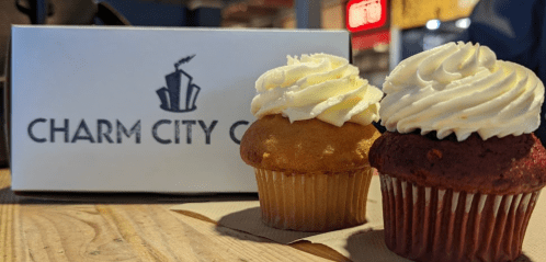 Two cupcakes, one vanilla and one red velvet, sit beside a box labeled "Charm City Cakes."