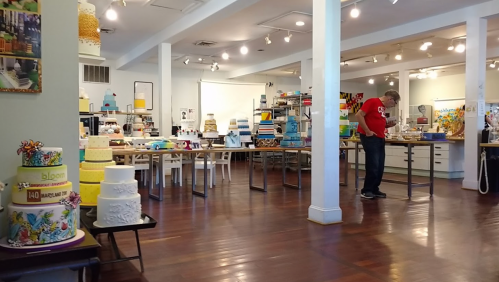 A spacious cake studio with various decorated cakes and a person examining the displays. Brightly lit and organized.