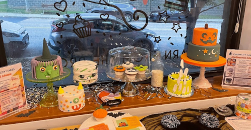 A display of colorful Halloween-themed cakes and cupcakes in a bakery window, with festive decorations around.