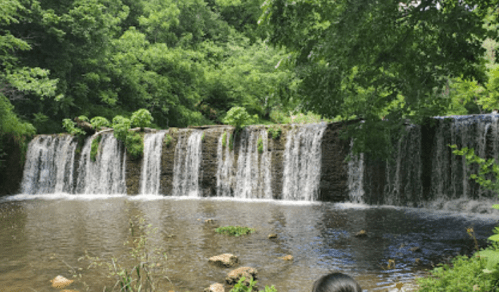 A serene waterfall cascading over rocks, surrounded by lush green trees and a calm pool of water below.