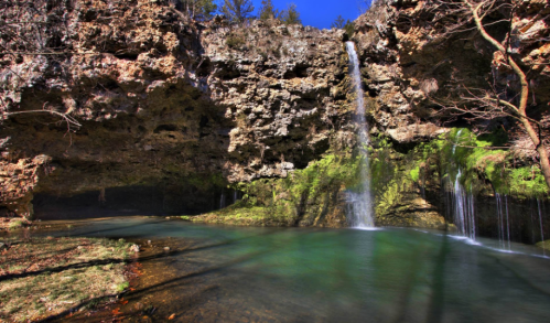 A serene waterfall cascades into a clear pool, surrounded by rocky cliffs and lush greenery under a bright blue sky.