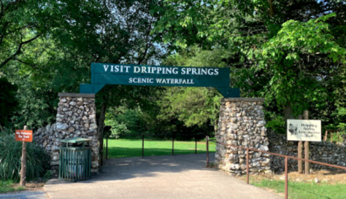 Sign at the entrance to Dripping Springs Scenic Waterfall, framed by stone pillars and surrounded by greenery.