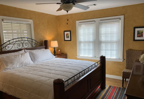 Cozy bedroom with a large bed, warm yellow walls, and natural light from two windows. A dresser and chair are visible.