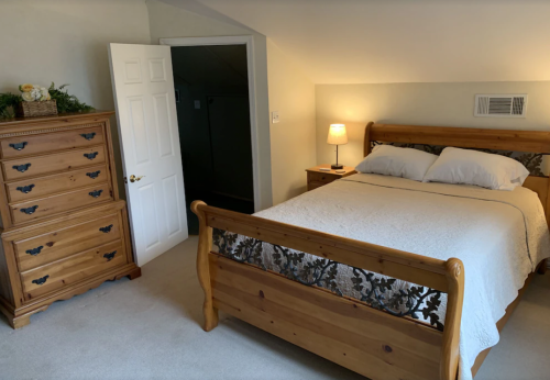 Cozy bedroom with a wooden bed, dresser, lamp, and a door leading to another room. Soft lighting and neutral decor.