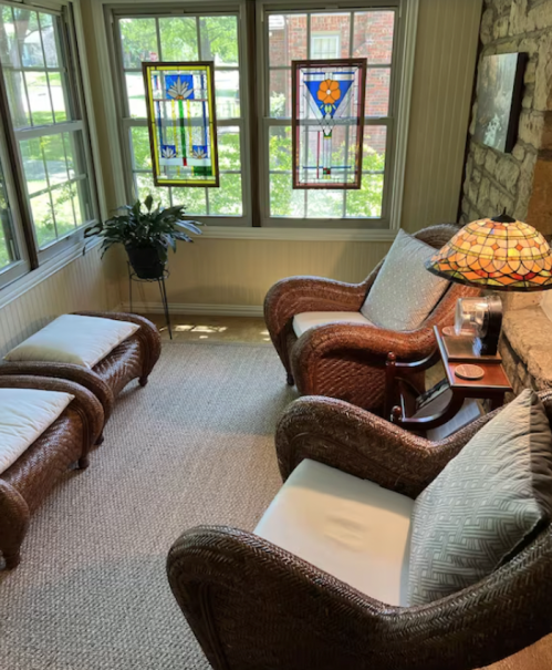 Cozy sunroom with two wicker chairs, a small table, and stained glass windows, surrounded by greenery.