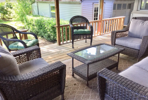 A cozy outdoor seating area with wicker furniture and a glass table on a wooden deck surrounded by greenery.