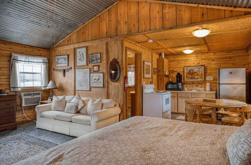 Cozy cabin interior featuring a sofa, dining area, kitchen, and rustic wooden walls. Warm and inviting atmosphere.