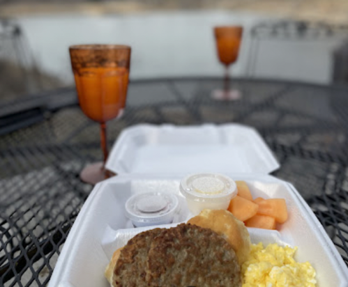A takeout breakfast with sausage, scrambled eggs, a biscuit, fruit, and drinks on a table by a lake.