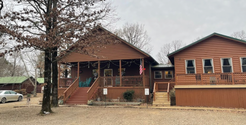 A rustic wooden cabin with a porch, surrounded by trees and gravel, alongside another similar building.