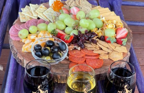A wooden platter filled with assorted cheeses, meats, fruits, crackers, and three glasses of wine.