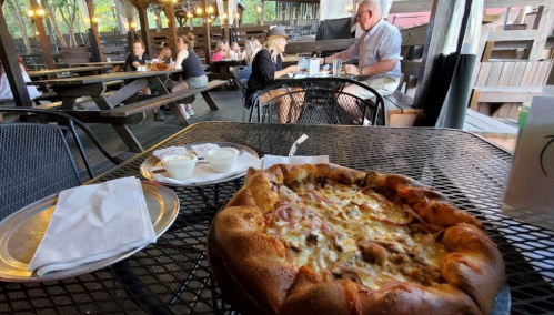 A table with a large pizza and dipping sauces, surrounded by diners in a rustic outdoor restaurant setting.