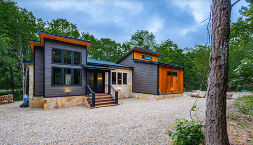 Modern two-story house with a mix of wood and stone exterior, surrounded by trees and a gravel driveway.