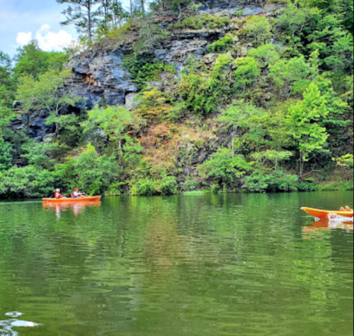 Two kayakers paddle on a calm lake surrounded by lush greenery and rocky cliffs.