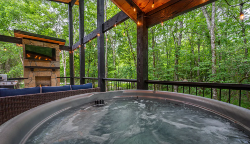 A hot tub on a deck surrounded by lush greenery, with string lights and a stone fireplace in the background.