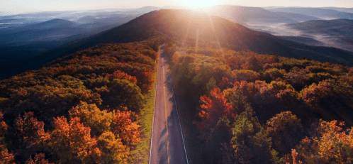Aerial view of a winding road through vibrant autumn foliage, with mountains and a sunrise in the background.