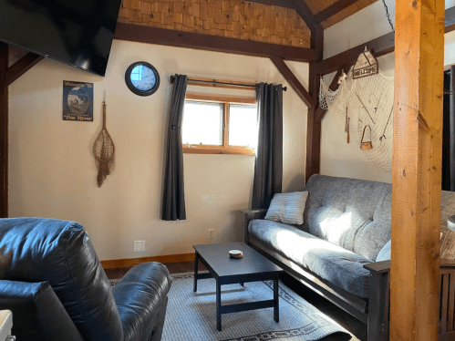 Cozy living room with a gray sofa, wooden beams, a clock, and nautical decor by a window with dark curtains.