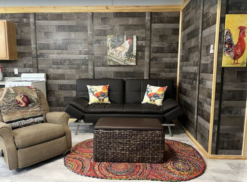 Cozy living room with a black sofa, colorful cushions, a woven coffee table, and rustic wooden walls.
