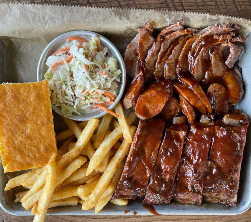 A tray of barbecue featuring sliced brisket, sausage, coleslaw, fries, and a piece of cornbread.