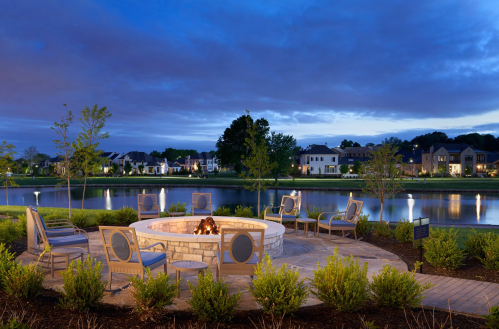 A serene lakeside fire pit area with seating, surrounded by greenery and homes, under a twilight sky.