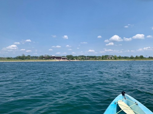 A view of calm water with a distant shoreline and blue sky dotted with clouds, seen from a kayak.