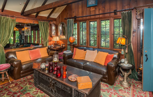 Cozy living room with wooden walls, two brown sofas, colorful cushions, vintage decor, and large windows.