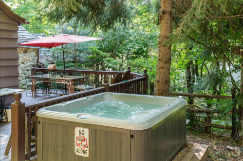 A hot tub on a wooden deck surrounded by trees, with a patio umbrella and outdoor seating nearby.