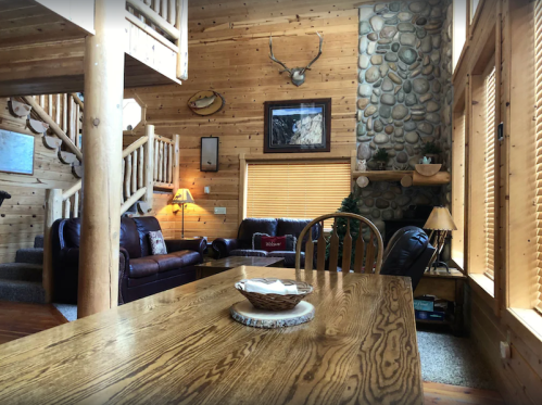 Cozy wooden cabin interior with a dining table, leather sofas, stone fireplace, and rustic decor.