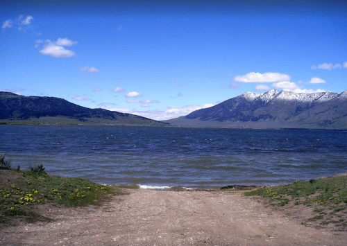A serene lake surrounded by mountains, with a clear blue sky and scattered clouds. Green grass and wildflowers in the foreground.