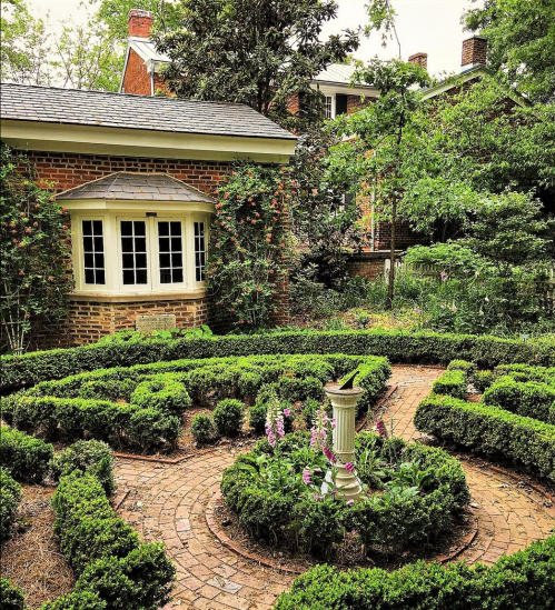 A lush garden with neatly trimmed hedges, a stone path, and a central pedestal surrounded by colorful flowers.
