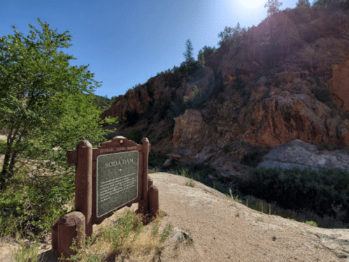 A historical sign near a rocky canyon, surrounded by trees and sunlight.