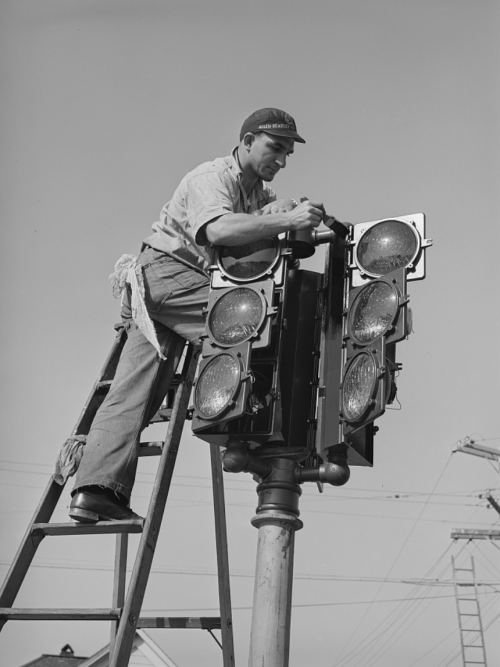 A worker on a ladder adjusts a traffic signal with multiple lights against a clear sky.