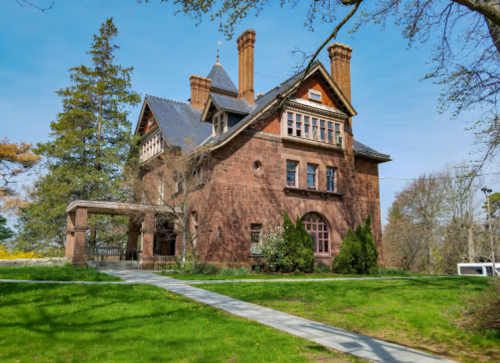 A large, historic brick mansion with a slate roof, surrounded by green grass and trees under a clear blue sky.