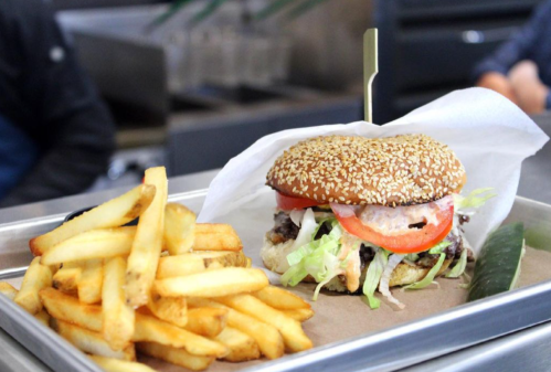 A sesame seed burger with lettuce, tomato, and sauce, served with a side of golden French fries on a metal tray.
