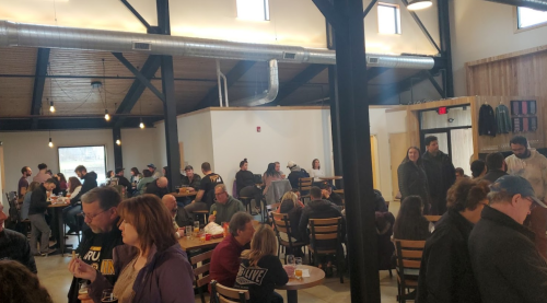 A lively indoor gathering with people socializing, seated at tables, and enjoying drinks in a spacious venue.