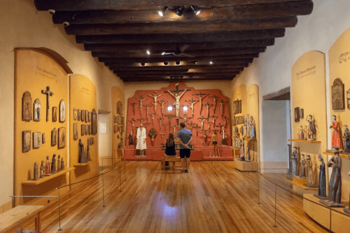 A museum interior featuring wooden sculptures and crosses on the walls, with a visitor observing the exhibits.