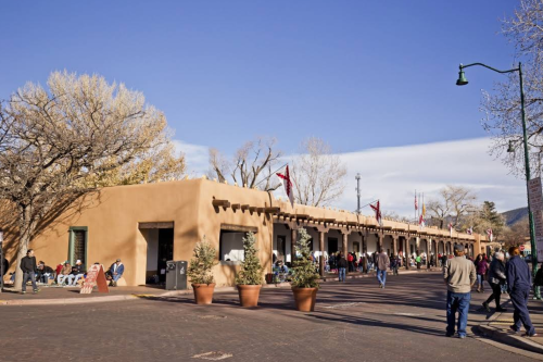 A bustling outdoor market with adobe-style buildings, trees, and people walking under a clear blue sky.