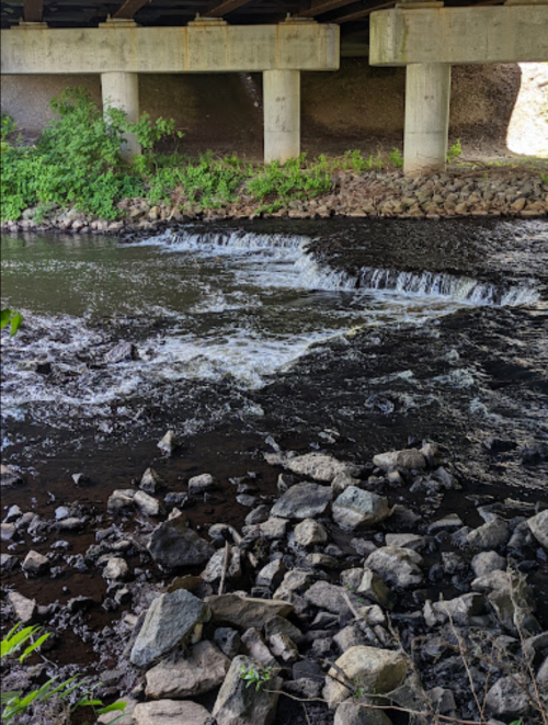A small waterfall flows under a bridge, surrounded by rocks and greenery along the riverbank.