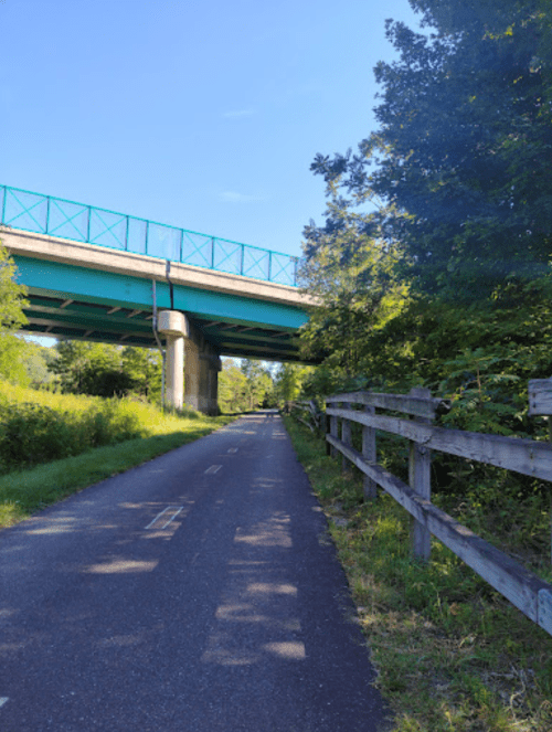 A paved path runs alongside a wooden fence, with a highway overpass and trees in the background under a clear blue sky.