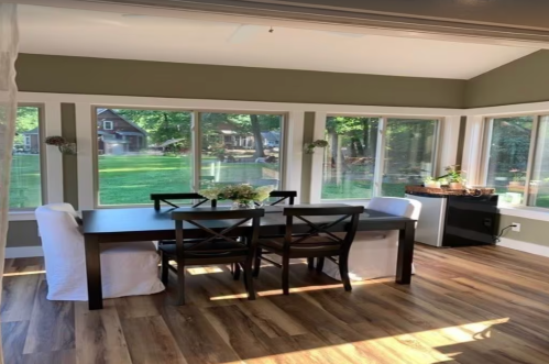 Bright dining area with a wooden table, chairs, and large windows overlooking a green yard.
