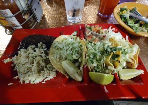 A red plate with three tacos topped with lettuce and cilantro, served with rice, black beans, pico de gallo, and lime wedges.