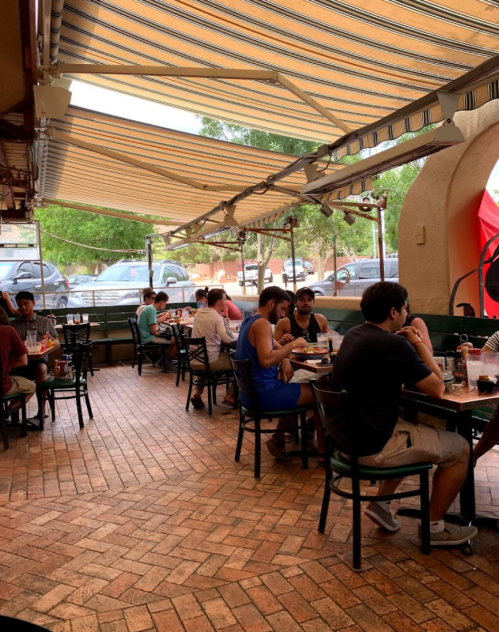 A bustling restaurant interior with diners at tables, large windows, and striped awnings letting in natural light.