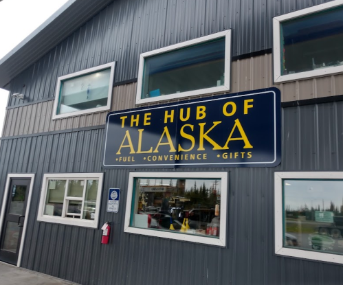 Sign for "The Hub of Alaska" on a building, featuring fuel, convenience items, and gifts. Windows visible.
