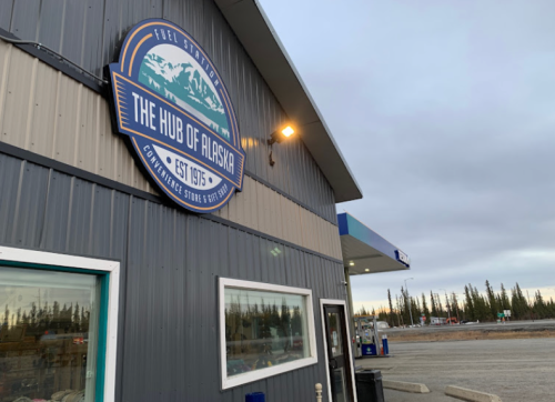 Sign for "The Hub of Alaska" on a building, with a fuel station and trees in the background under a cloudy sky.