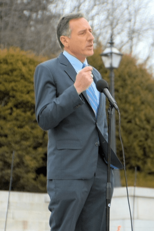 A man in a suit speaks at a microphone outdoors, with trees and a lamp post in the background.